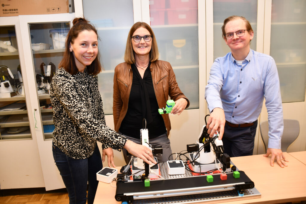 Fachlehrerin Franziska Grimes, Gesamtschuldirektorin Dr. Petra Hoppe und Fachlehrer Julian Frankl stehen vor einem Roboter.