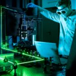 Student mit Schutzbrille steht in einem abgedunkelten Labor und justiert einen grünen Laser auf einem Tischaufbau und hat vor sich einen Laptop stehen.