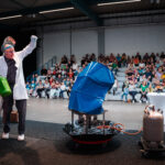 Physiker auf einer Bühne vor einem jungen Publikum mit einem blauen implodierten Fass
