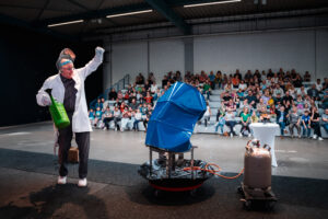 Physiker auf einer Bühne vor einem jungen Publikum mit einem blauen implodierten Fass